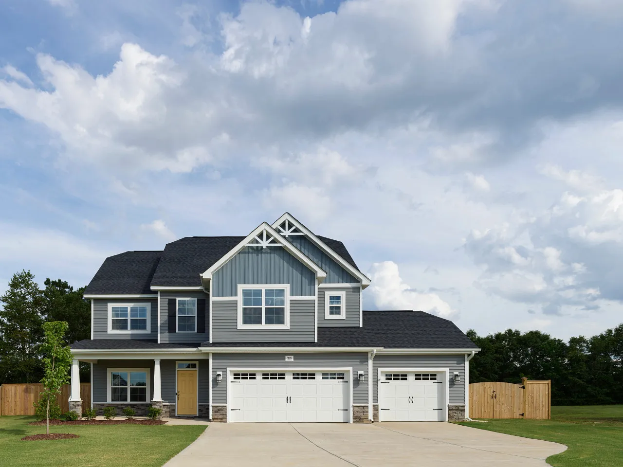 Two-story gray house with white trim, a triple garage, and a large driveway under a partly cloudy sky.